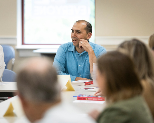 A smiling man participates in a book club discussion at the Lexington Public Library. He sits at a table with several other adults, surrounded by books, name cards, and pens. The mood is friendly and engaged, with blurred participants in the foreground having lively conversation. 